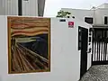 Frontale Farbfotografie von einer weißen Mauer, an die „Der Schrei“ gemalt wurde. Rechts sind eine kleine Tür und ein Tor. Im Hintergrund sind Gebäuden zu erkennen. Beim abgebildeten Gemälde fehlt der schreiende Mensch.