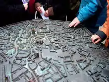 Blick von schräg oben auf ein bronzenes Relief, das modellhaft die Altstadt von Münster zeigt. Es ist in der Fußgängerzone der Münsteraner Altstadt etwa in Hüfthöhe horizontal auf einen Sockel montiert. Von den dicht dahinter stehenden Passanten sieht man nur die Hüften, einige betasten mit ihren Händen die Details des Modells. Dieses zeigt leicht stilisiert Straßen, Plätze, Gebäude und Baumgruppen; die Häuser sind etwa 2 cm hoch. Der Dom, die Lamberti- und die Liebfrauenkirche ragen mit mehreren Zentimetern Höhe deutlich heraus. Die Straßen sind in Brailleschrift mit ihren Namen gekennzeichnet. Am rechten Bildrand erkennt man Teile einer Widmung; dieser mehrzeilige Text ist sowohl in Brailleschrift als auch in erhabener Normalschrift ausgeführt.