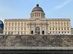 Farbfotografie in der Untersicht von einem barocken Nachbau mit Ufermauer im Vordergrund. Das dreigeschossige, helle Gebäude hat in der Mitte Säulen und Ornamente. Auf dem Dach befindet sich eine Kuppel mit goldenem Kreuz.