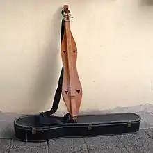 Ein längliches an eine Wand gelehntes Dulcimer. Das Instrument ist eine leicht geschwungene Kastenzithern mit flachem Korpus. Das Griffbrett ist direkt aufgesetzt. In den Korpus sind vier herzförmige Öffnungen eingelassen.