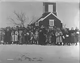 Schwarz-Weiß-Foto einer Gruppe von in Pelzmänteln gekleideter Personen vor einer Holzkirche