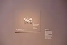 Braungraue Wand, vor der ein Rechteck in derselben Farbe in Größe des verliehenen impressionistischen Bildes hängt. Darauf ist klein mittig das verliehene Bild in Grautönen gedruckt, daneben Text auf Deutsch und Englisch