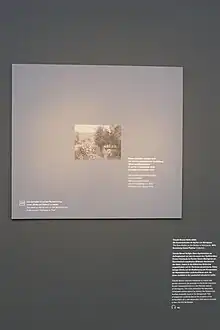 Braungraue Wand, vor der ein Rechteck in derselben Farbe in Größe des verliehenen impressionistischen Bildes hängt. Darauf ist klein mittig das verliehene Bild in Grautönen gedruckt, daneben Text auf Deutsch und Englisch