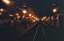 Dunkles Foto eines unterirdischen, mit Glühlampen schwach erleuchteten Bahnhofs. Blick entlang einer Gleisachse, die glänzenden Schienenköpfe laufen in die Ferne, rechts und links sind Bahnsteige mit charakteristischen Stützenreihen in der relativ starken Dunkelheit auszumachen.