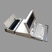 Notebook. Kastenförmiges Gehäuse aus beigem Kunststoff, hinter der Tastatur die Schlitze zweier Diskettenlaufwerke, dahinter hochgeklappt ein schmaler Bildschirm. Hinten angedockt ein schmaler Drucker derselben Bauhöhe.
